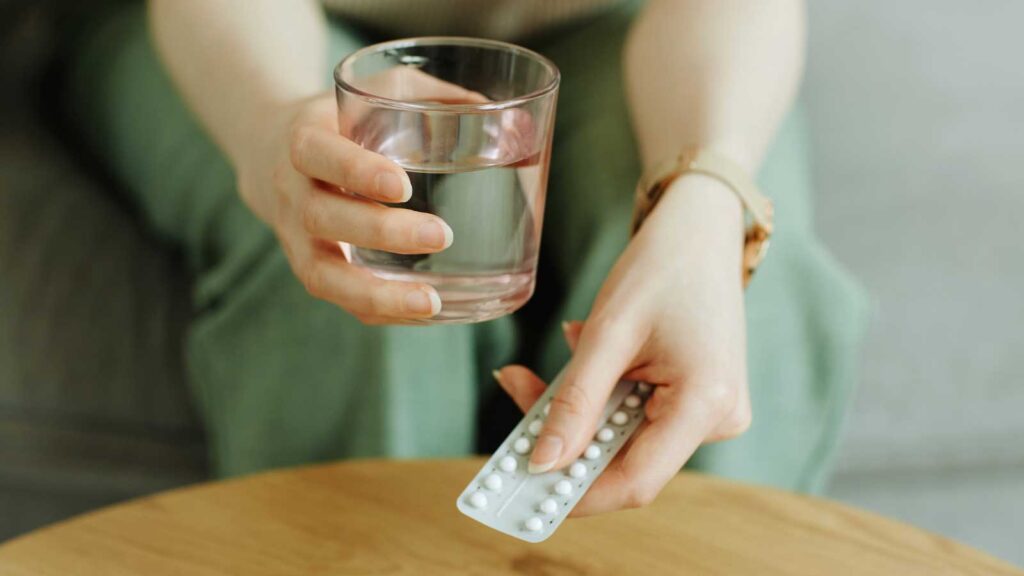 A person's hands holding a glass of water and a white blister pack of birth control pills, positioned over a light wooden table, with a soft, blurred background. 