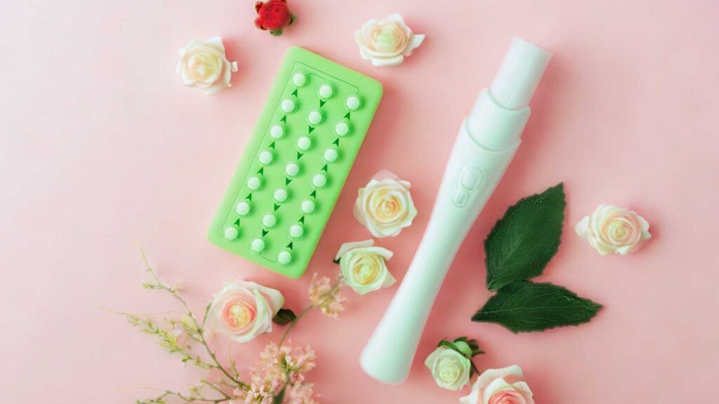 A top-down view of a light green blister pack of birth control pills and a white, pen-shaped vaginal applicator, surrounded by scattered white and pink roses and green leaves on a soft pink background. 